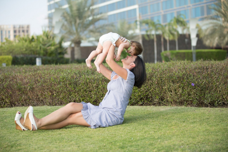 Eine junge Mutter sitzt auf dem Rasen und hält ihr Baby hoch. 